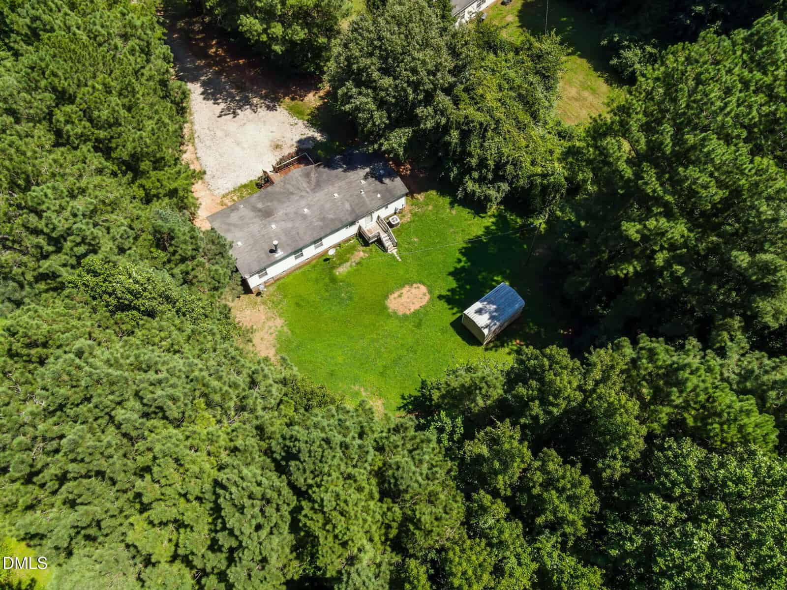 Aerial view of a single-story house surrounded by dense trees, with a grassy yard, small shed, and gravel driveway.
