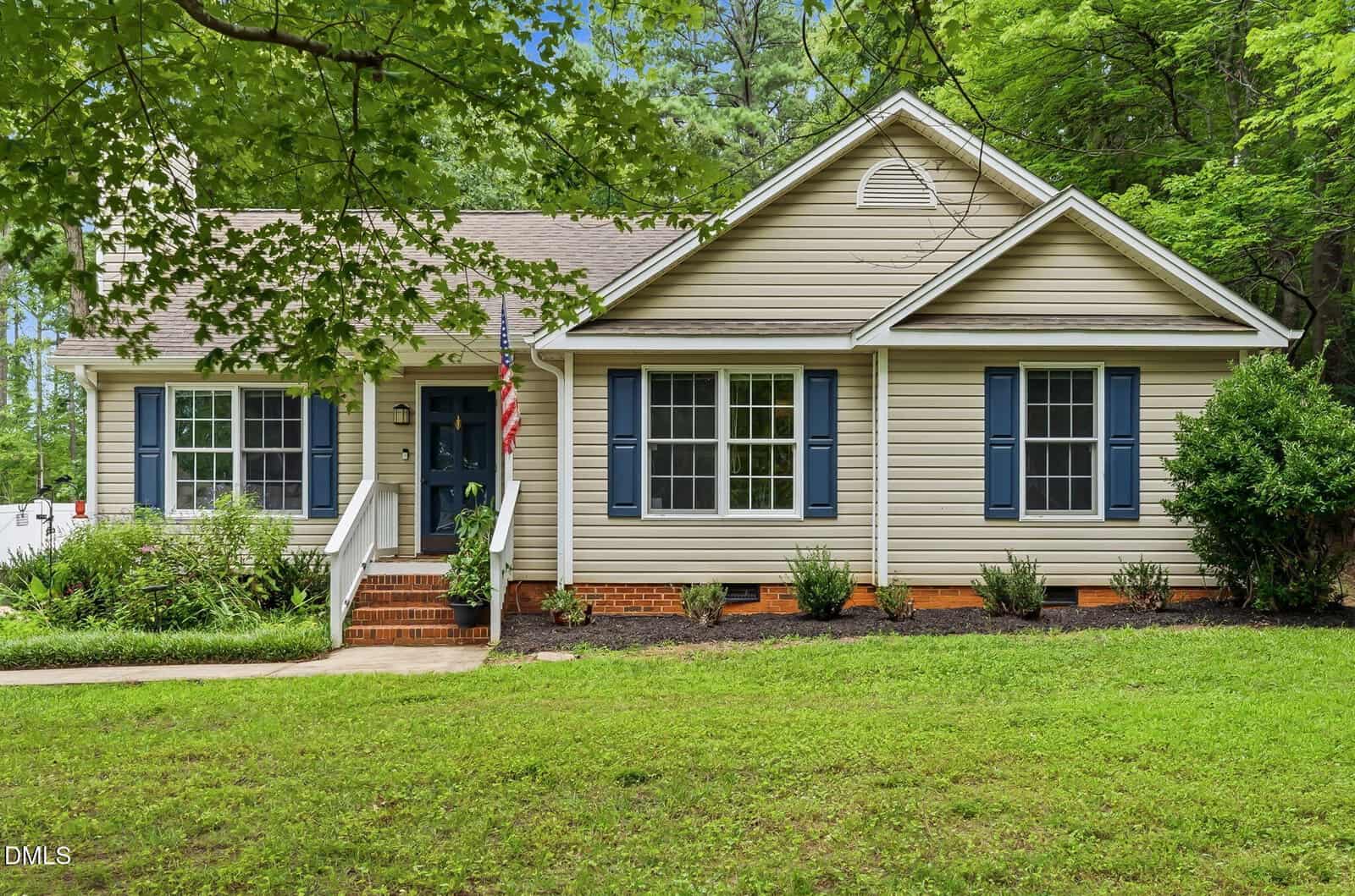 Charming single-story house at 114 Black Cloud Drive, Louisburg, featuring beige siding, blue shutters, and a classic gable roof. There are three front windows, a front door with steps and railing, a patch of lawn, and mature trees surrounding the property.
