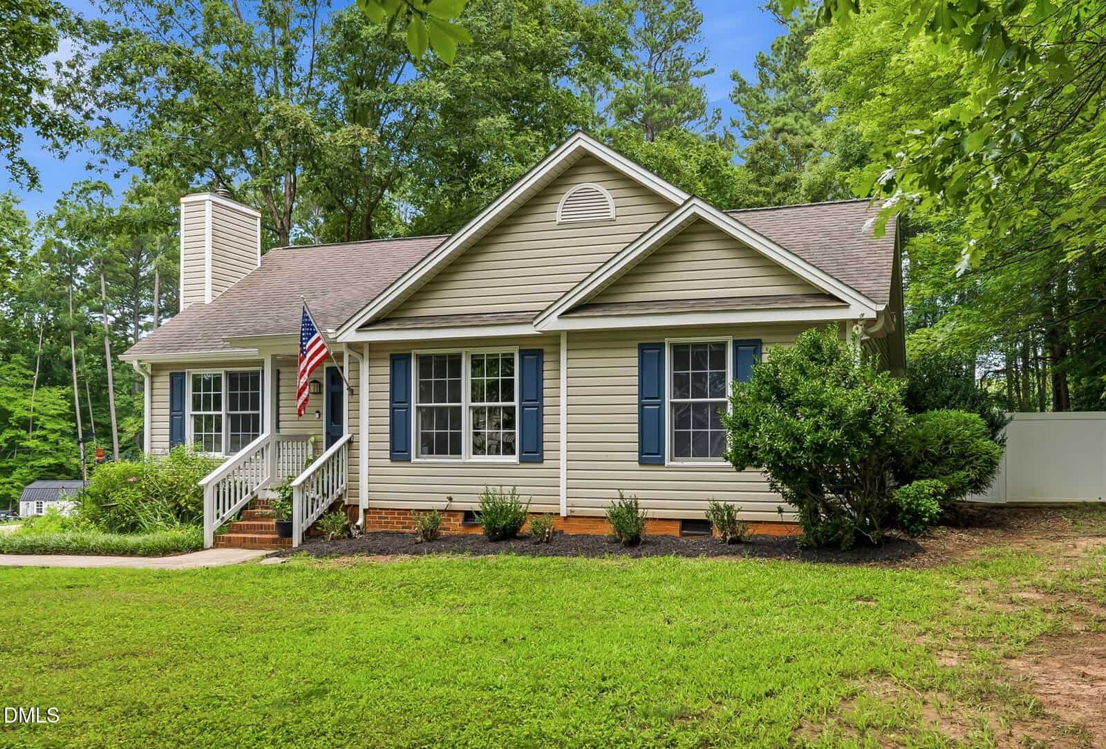 Single-story beige house with blue shutters, a gable roof, front steps with white railings, an American flag, and landscaped bushes. Lush green lawn and trees surround the home under a clear sky.