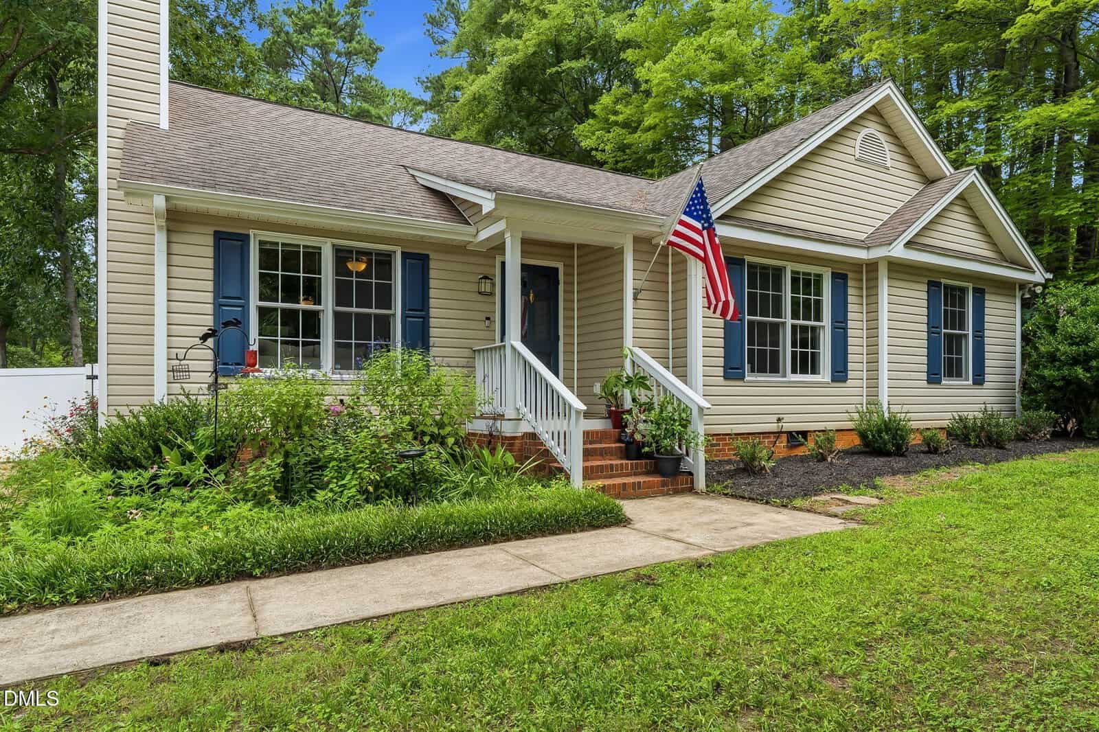 A beige single-story house with blue shutters, a covered porch, an American flag, and a landscaped front yard with green grass and shrubs. Tall trees are in the background.