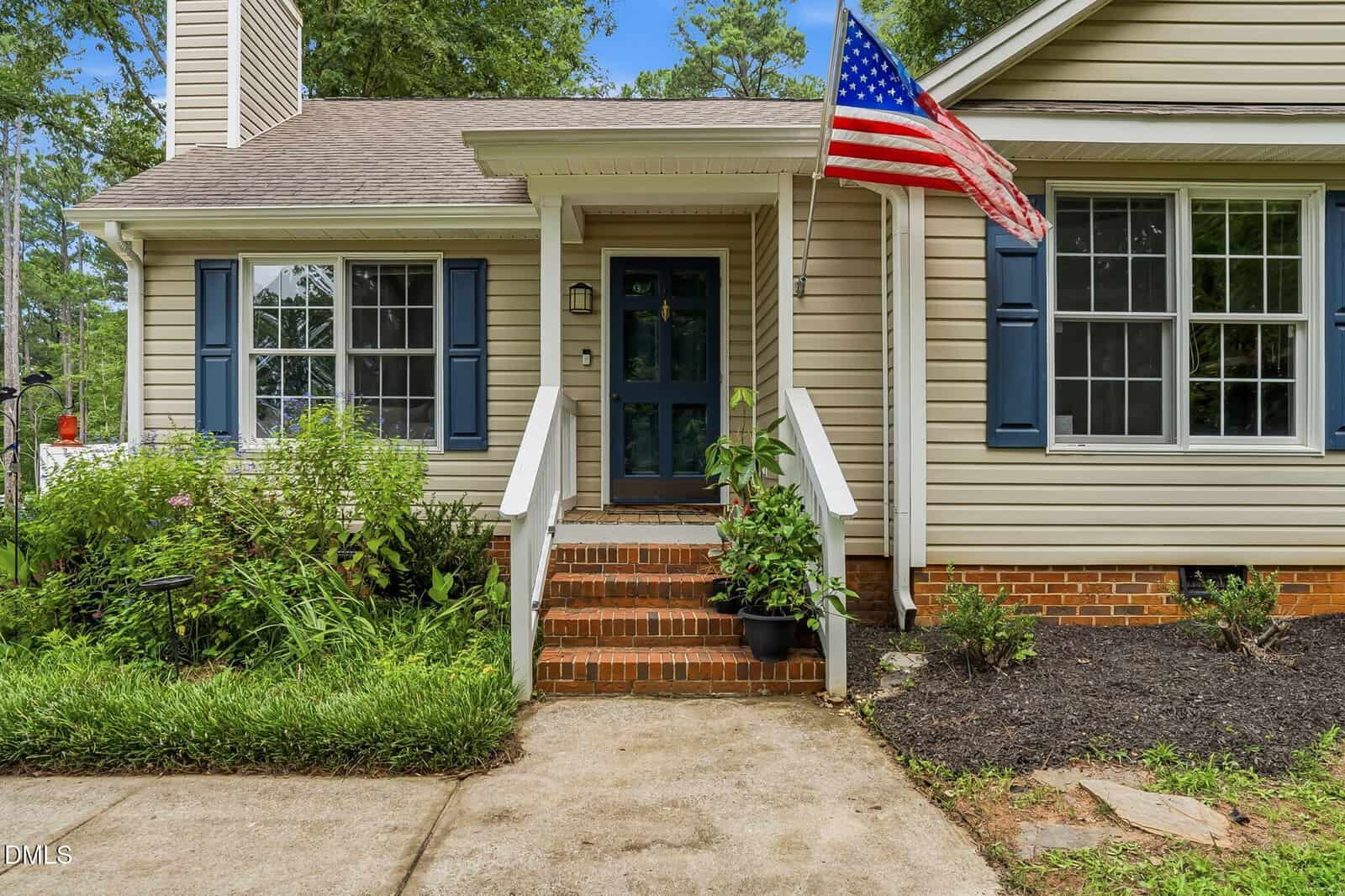 Front porch of a beige house with blue shutters, a blue door, brick steps, white railings, an American flag, potted plants, and surrounding greenery. A sidewalk leads up to the entrance.