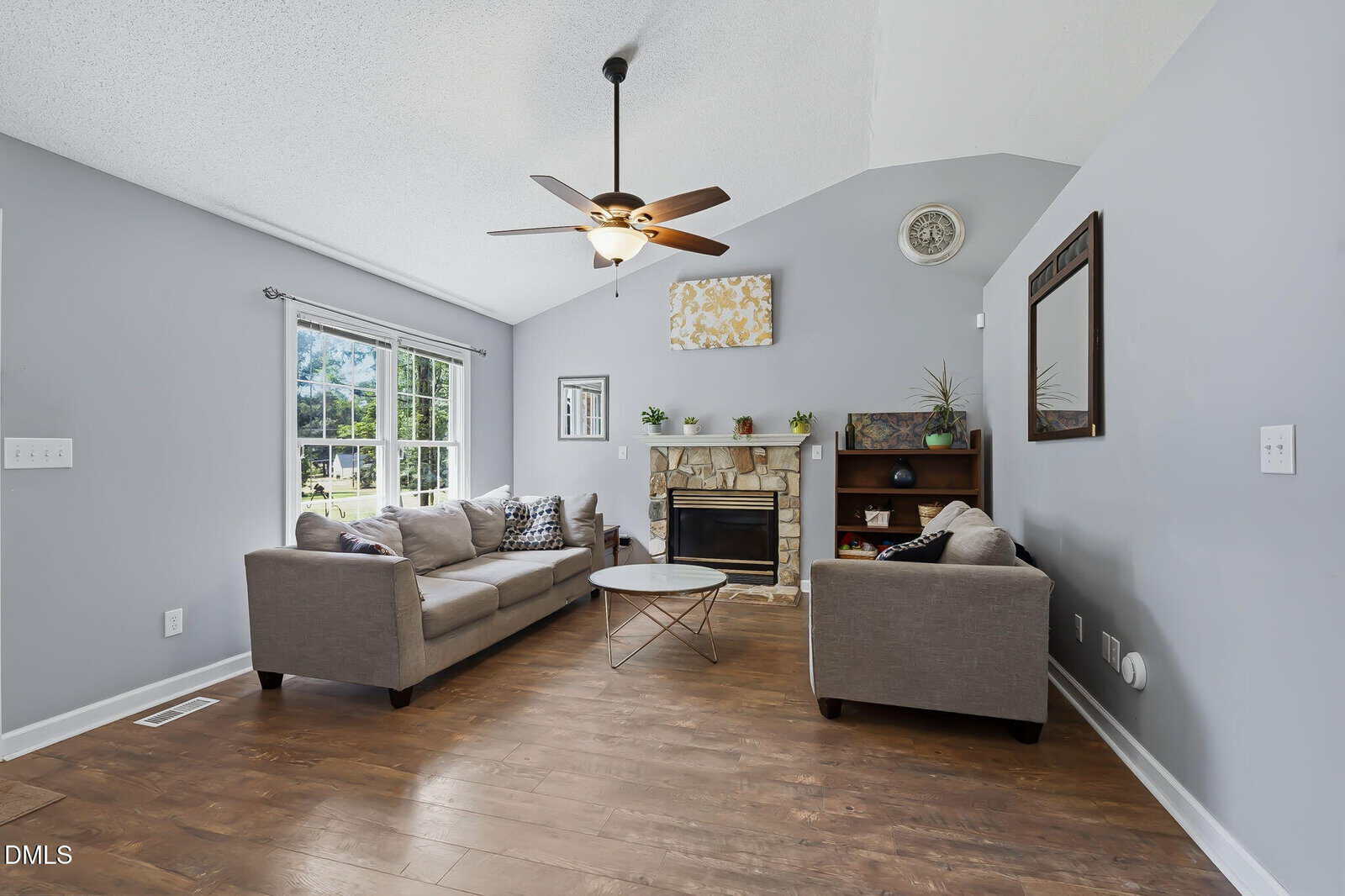Living room with vaulted ceiling, gray walls, wood floor, two gray sofas, round coffee table, stone fireplace, bookshelf, large window, ceiling fan, and wall decor.