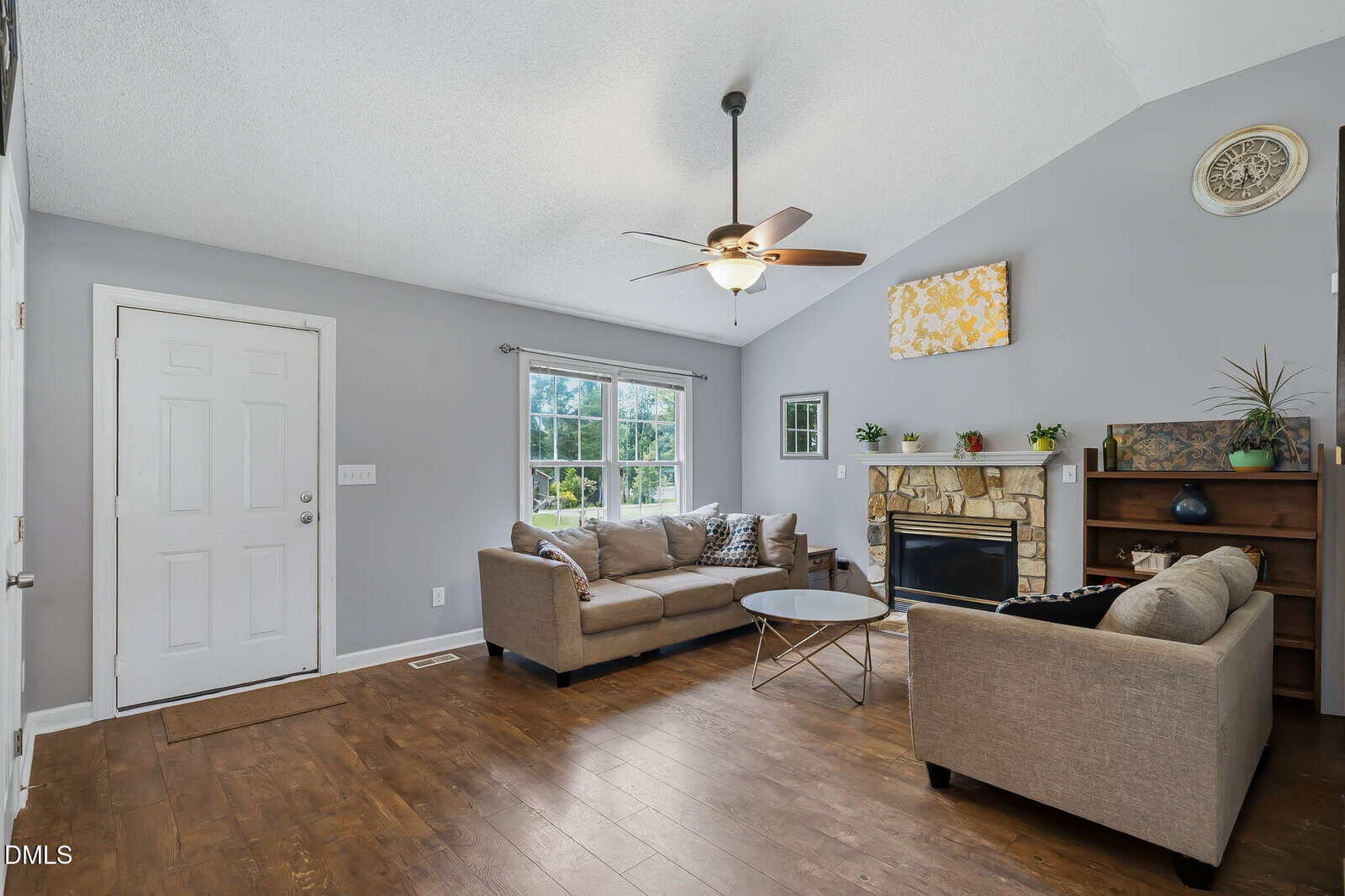 Living room with gray walls, wood floor, beige sofas, stone fireplace, plants, art above mantle, ceiling fan, and large window letting in natural light.