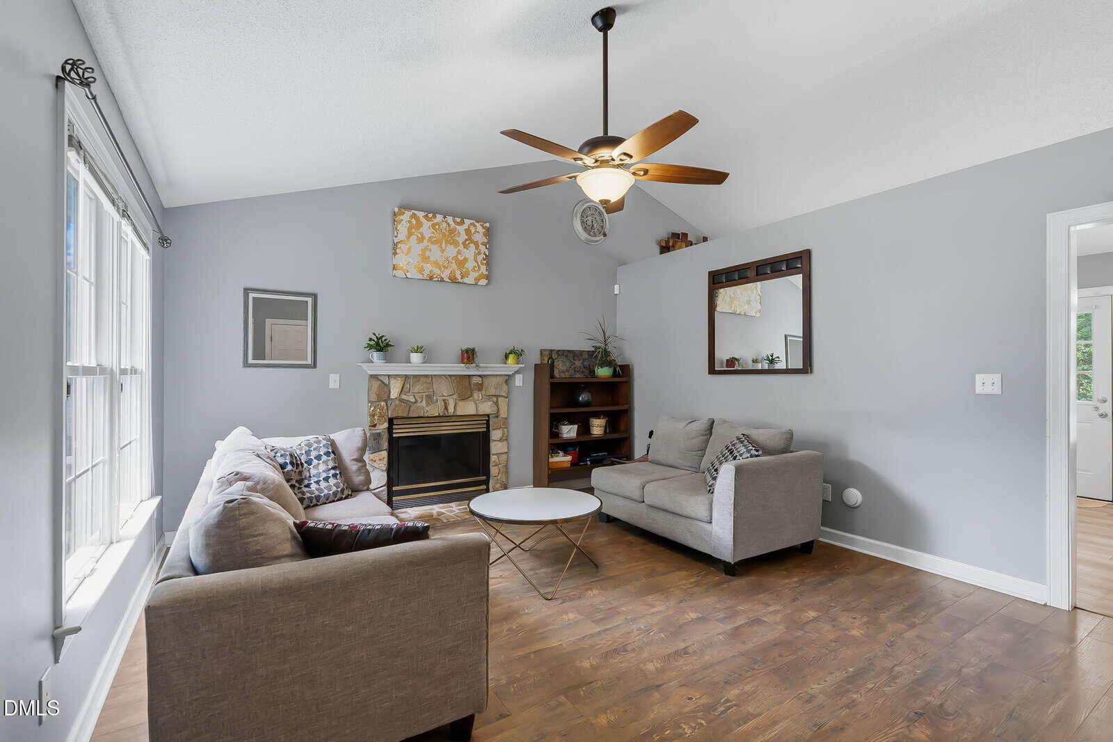 Modern living room with gray walls, hardwood floors, two sofas, a round coffee table, a fireplace with a mantel, shelves with decor, and a ceiling fan. Large window lets in natural light.