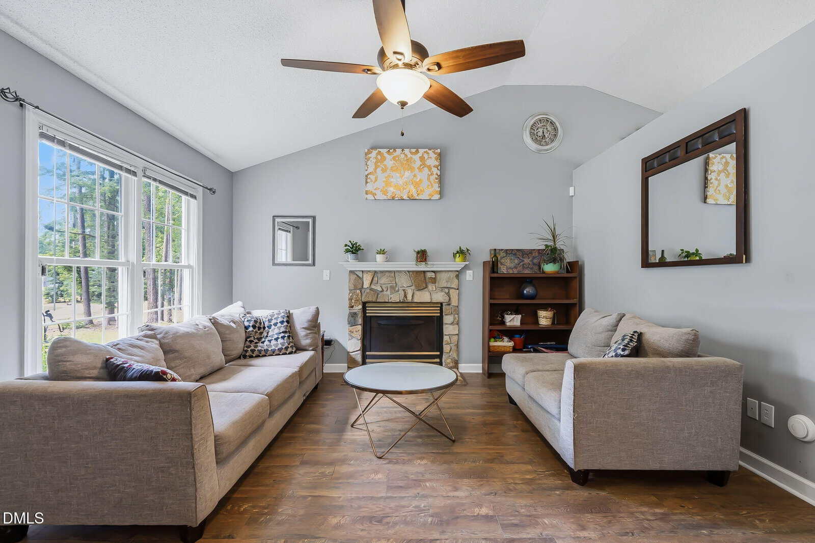 A living room with two gray sofas facing each other, a stone fireplace, a ceiling fan with light, two mirrors, a shelf with plants and decor, large windows on the left, and wood flooring.