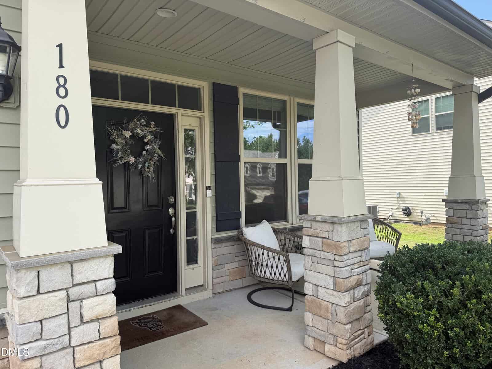 Front porch of a house at 180 Clubhouse Drive in Youngsville with a black door, a wreath, the number 180, two stone columns, a doormat, a chair with pillows, and a small shrub. Neighboring house and grass visible in the background.