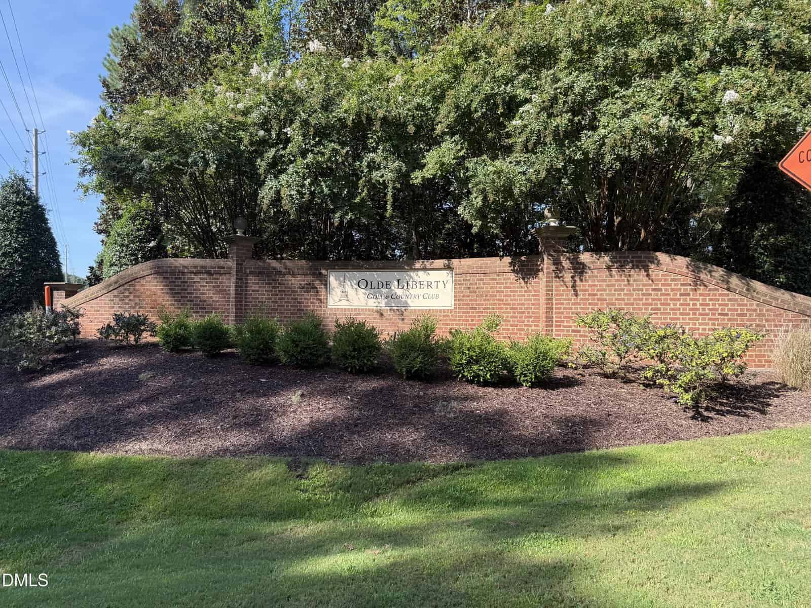A brick sign surrounded by bushes and trees reads “Olde Liberty Golf & Country Club” at 180 Clubhouse Drive, Youngsville, on a sunny day, with well-kept landscaping in front.
