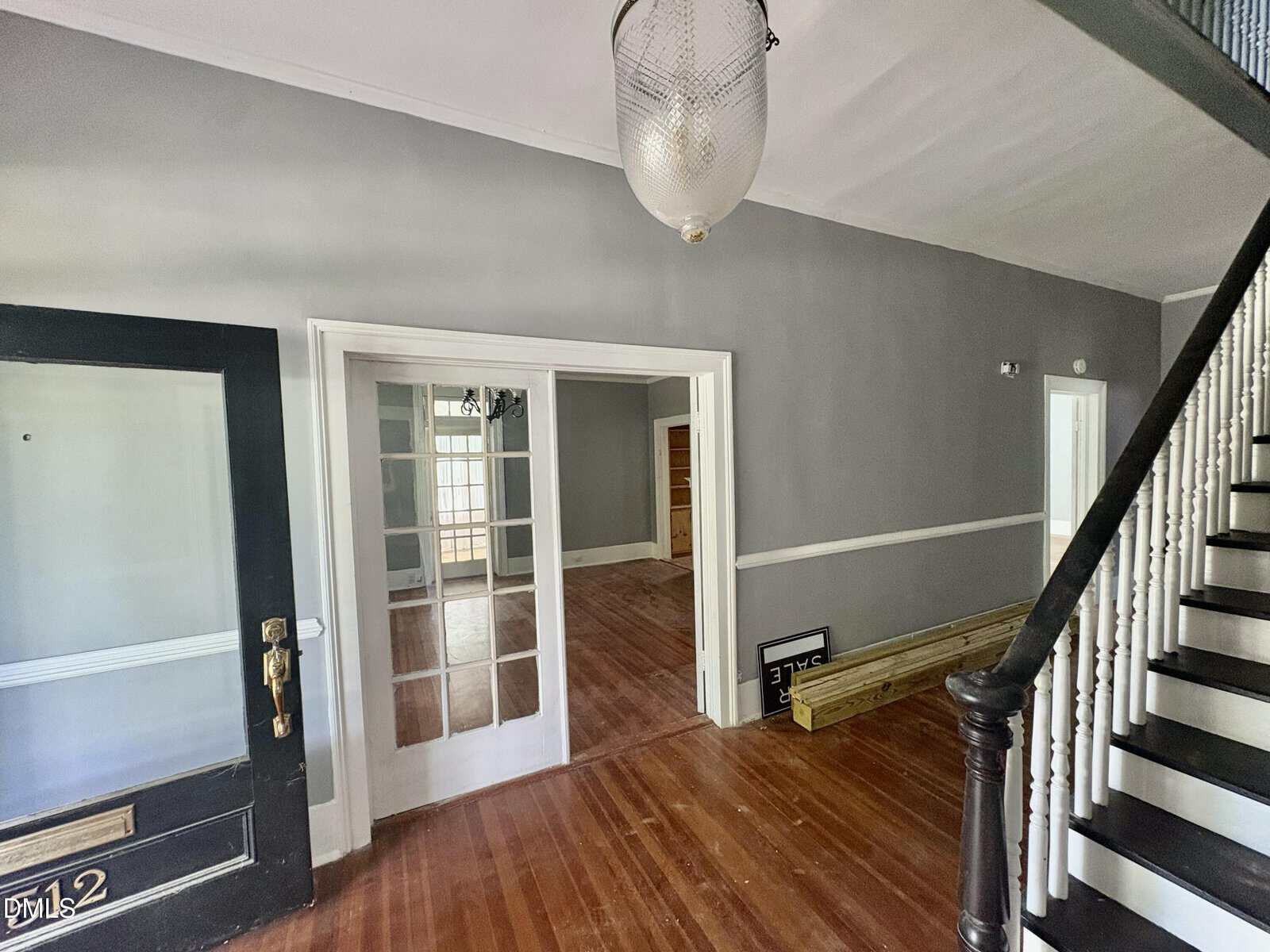 A hallway with hardwood floors, gray walls, a black staircase with white spindles, a glass door, and French doors leading to bright rooms. A light fixture hangs from the ceiling; wooden planks rest on the floor.