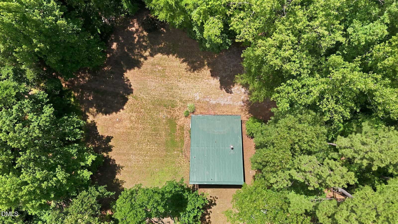 Aerial view of a small green-roofed building centered in a grassy clearing, surrounded by dense green trees on all sides. Shadows of trees are cast over parts of the grass and roof.