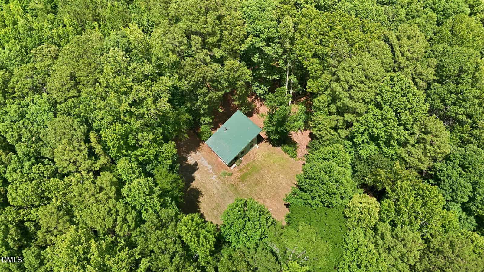 Aerial view of a small building with a green roof surrounded by dense green trees, with a clearing around the structure and a dirt path visible.