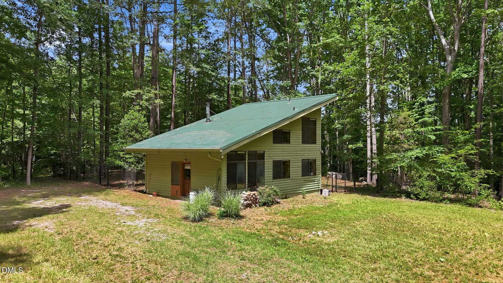 A small green house with a slanted roof sits in a grassy clearing surrounded by tall trees and dense forest.