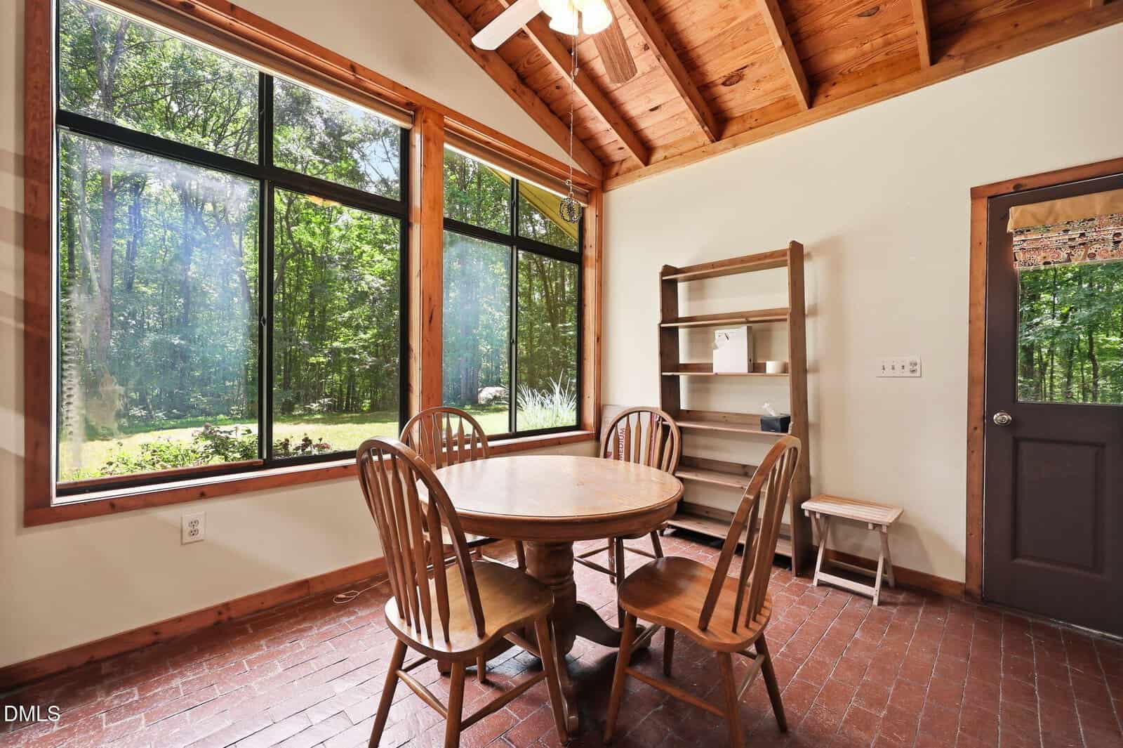 Dining area with round wooden table, four chairs, brick floor, a bookshelf, and large windows showing trees and greenery outside, under a vaulted wood ceiling with exposed beams.