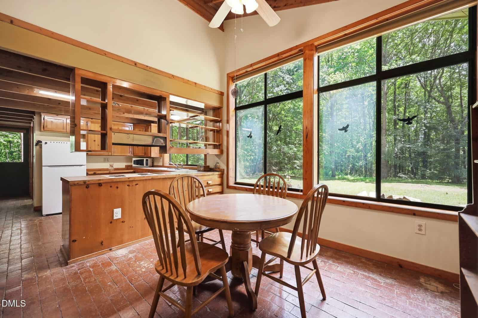 A cozy dining area with a round wooden table and four chairs sits by a large window overlooking a green yard. The open kitchen features wooden cabinets, shelves, and white appliances.