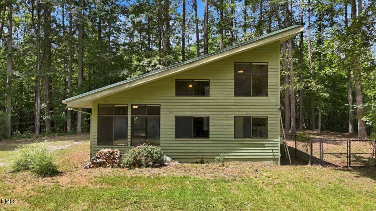 A green, modern house with a slanted roof sits surrounded by tall trees at 221 Allens Way. Large windows cover the front of the Kittrell home, and a small pile of wood is stacked near the foundation. There is a fenced area to the right.