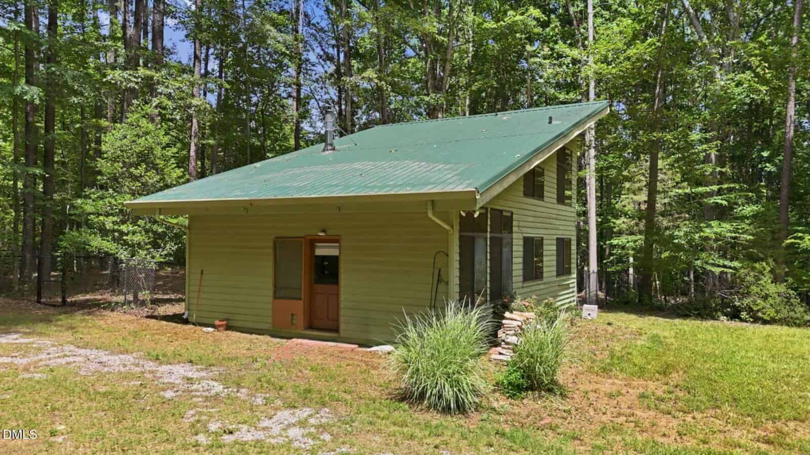 A small, green, two-story house with a sloped metal roof sits surrounded by tall trees and grass under a clear sky. There is a wooden door, several windows, and some plants near the entrance.