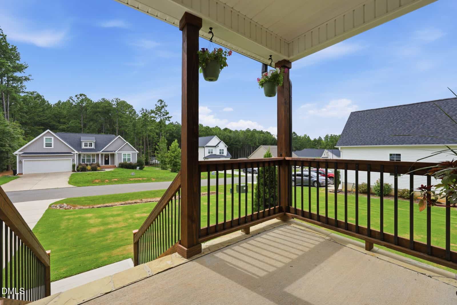 View from a covered front porch with wooden railings, looking out onto a suburban street with green lawns, neighboring houses, and trees in the background. Two hanging plants are attached to the porch ceiling.