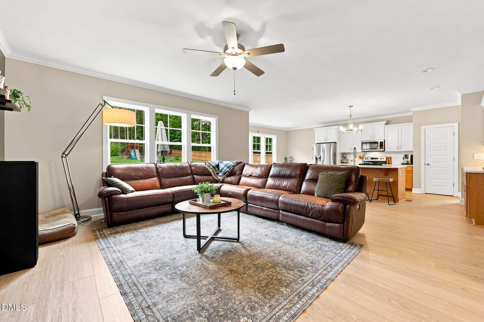 Spacious open-concept living room with a brown leather sectional sofa, round coffee table on a blue-patterned rug, ceiling fans, large windows, and a view of the kitchen and backyard playground.