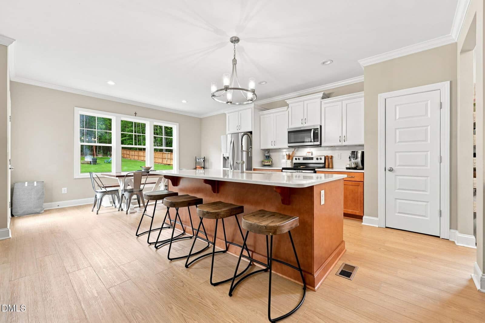 Modern kitchen with a large island, three wooden stools, white cabinets, stainless steel appliances, light wood flooring, and a dining area by large windows showing a backyard with green grass and a wooden fence.