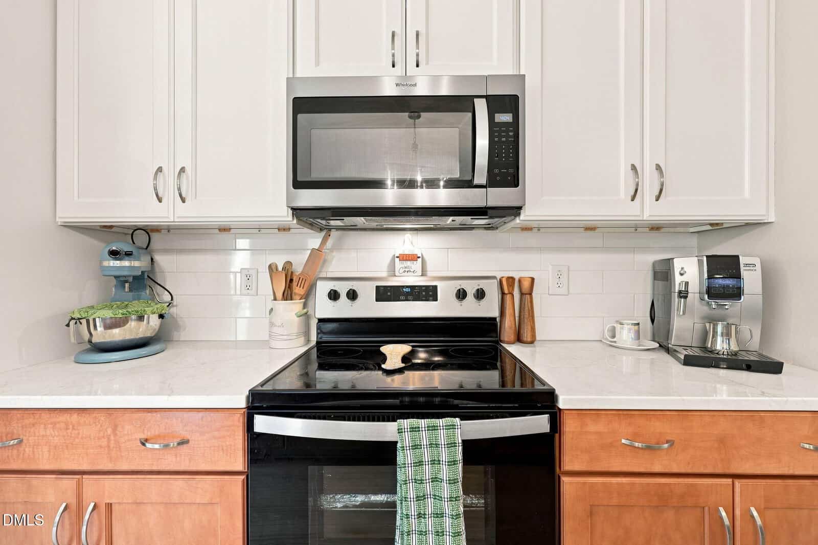 Modern kitchen with white cabinets, stainless steel microwave and oven, a stand mixer on the left, a coffee machine on the right, and cooking utensils plus a green towel hanging on the oven handle.