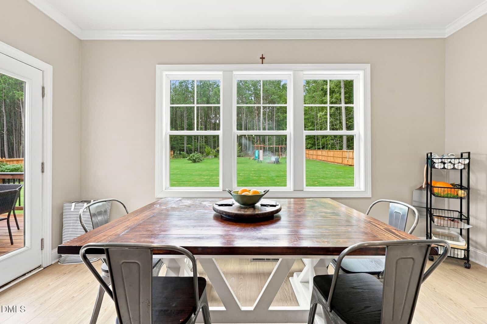 A bright dining area with a wooden table, four metal chairs, a bowl of fruit in the center, and large windows showing a green backyard and trees. A small rack with fruit sits in the corner by the window.