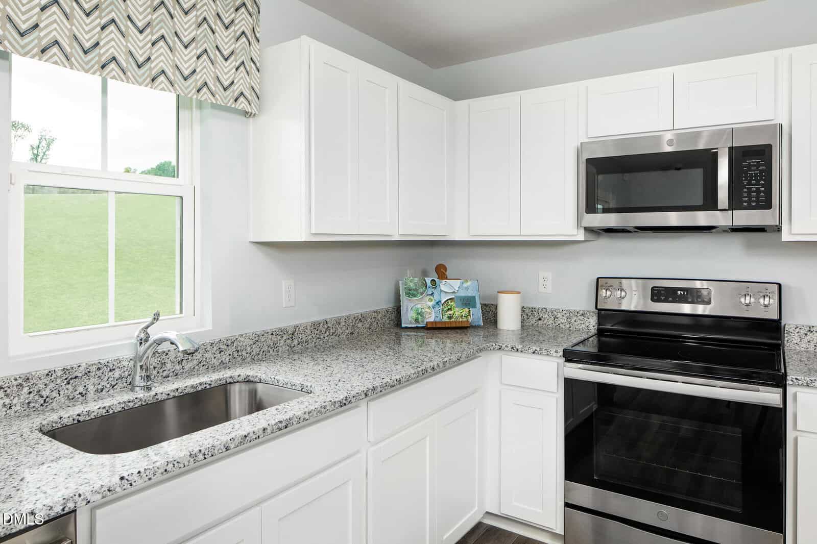 Modern kitchen with white cabinets, stainless steel appliances, granite countertops, a window with a patterned valance, and a sink. Cookbooks and a paper towel holder sit on the counter.