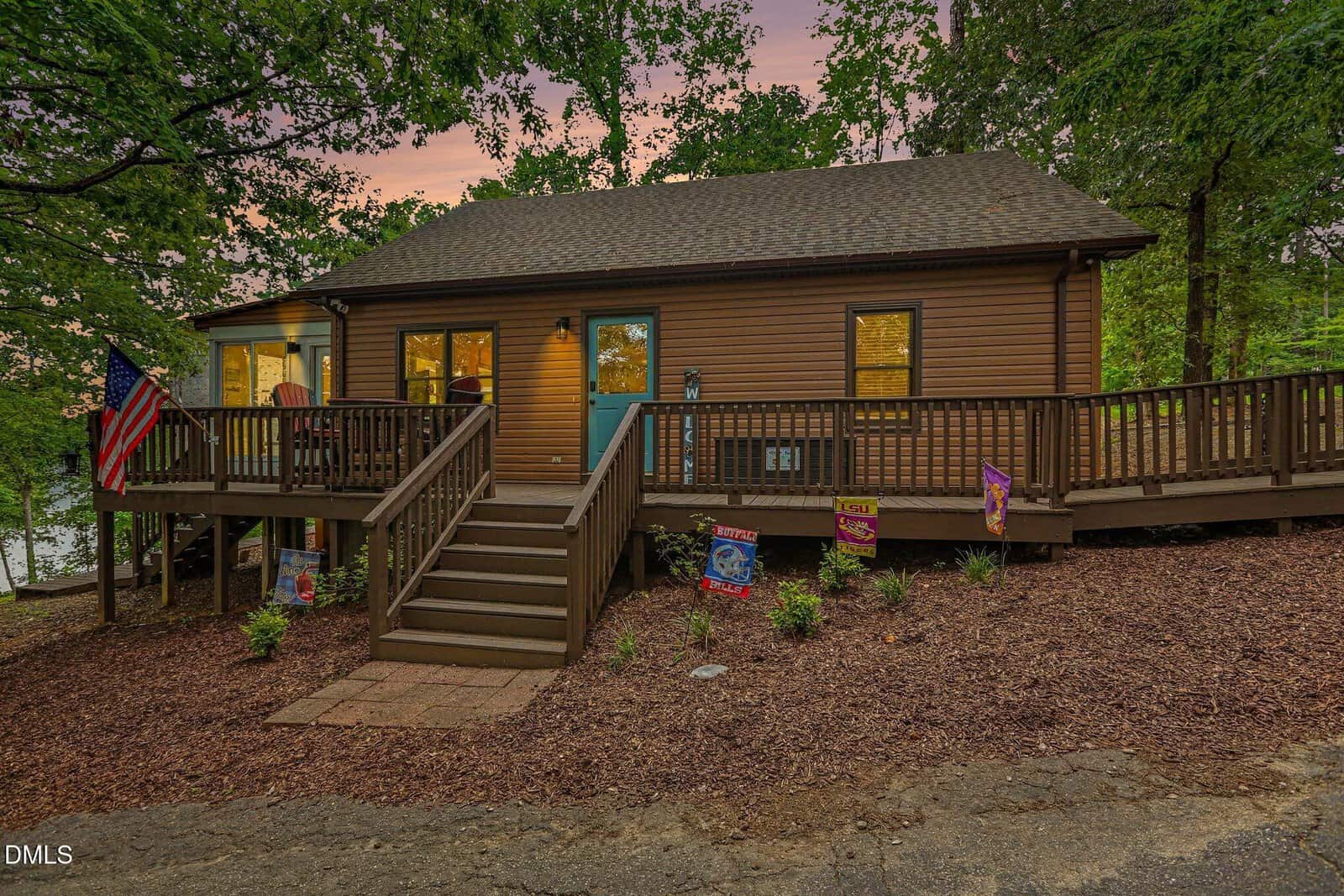 Charming brown single-story house at 207 Sagamore Drive, Louisburg, featuring a wooden deck and steps leading to a teal front door. Various flags are proudly displayed, and the property is surrounded by mature trees, with a lake partially visible in the background at sunset.