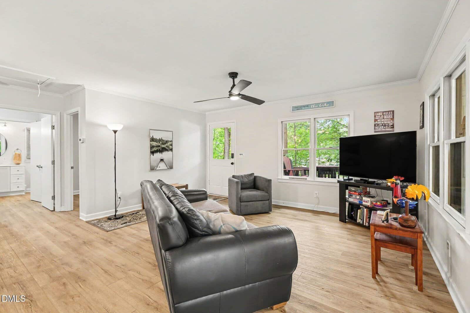 Living room with two gray sofas, a floor lamp, a TV on a stand, bookshelves, wood flooring, white walls, and large windows letting in natural light. A hallway leads to another room with a round mirror on the wall.