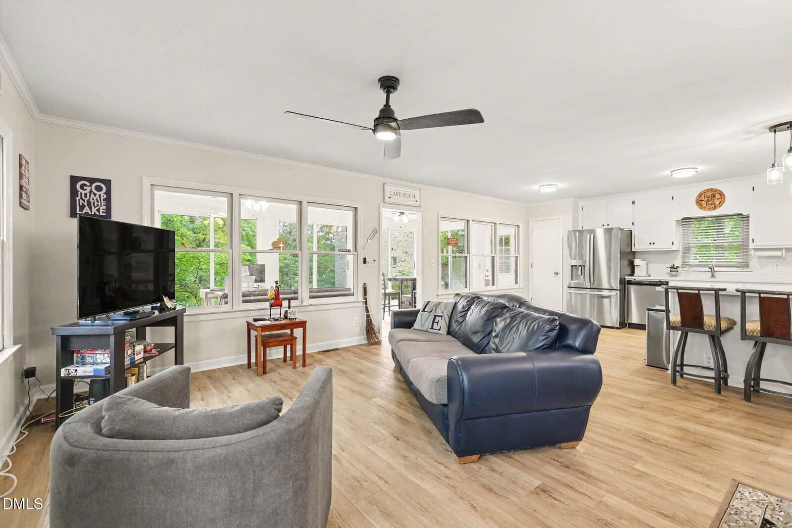Open-concept living area with wood flooring, a blue-gray sofa, a round gray chair, a wall-mounted TV, and a kitchen with white cabinets, stainless steel appliances, and an island with three stools. Large windows provide natural light.