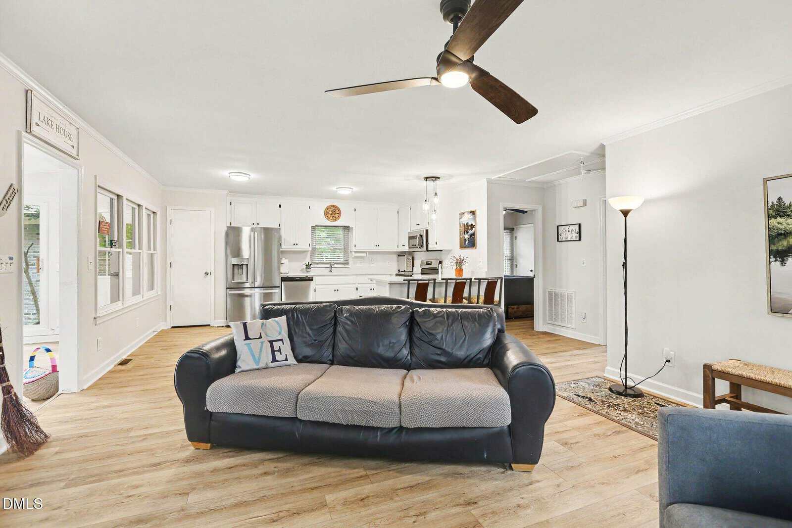 Open-concept living room and kitchen with a gray sofa, ceiling fan, floor lamp, wooden floors, white cabinets, stainless steel appliances, and a dining table with chairs. Bright and spacious space with modern decor.