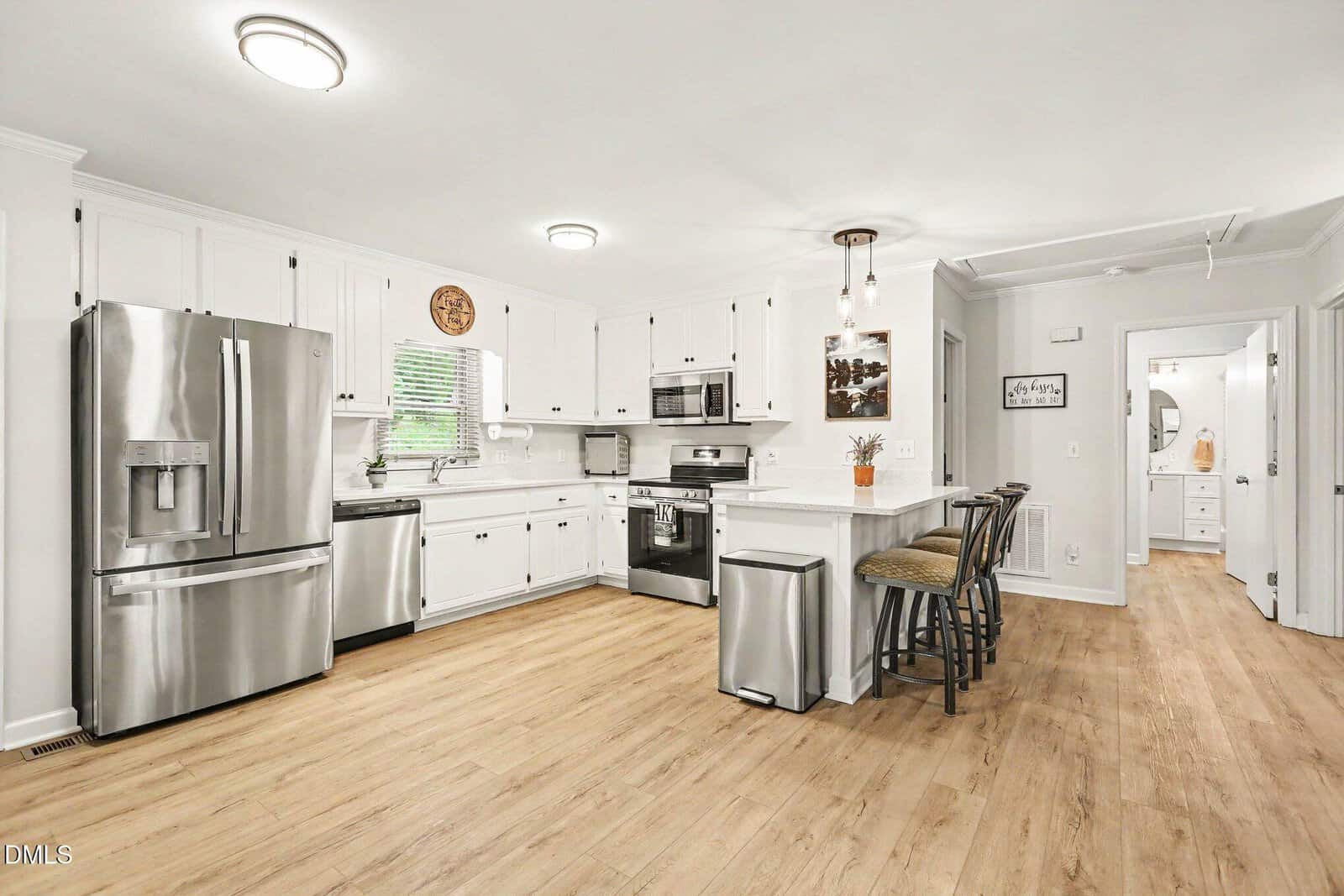 Modern kitchen with white cabinets, stainless steel appliances, light wood flooring, and a breakfast bar with three stools. There is bright overhead lighting and a hallway leading to other rooms.