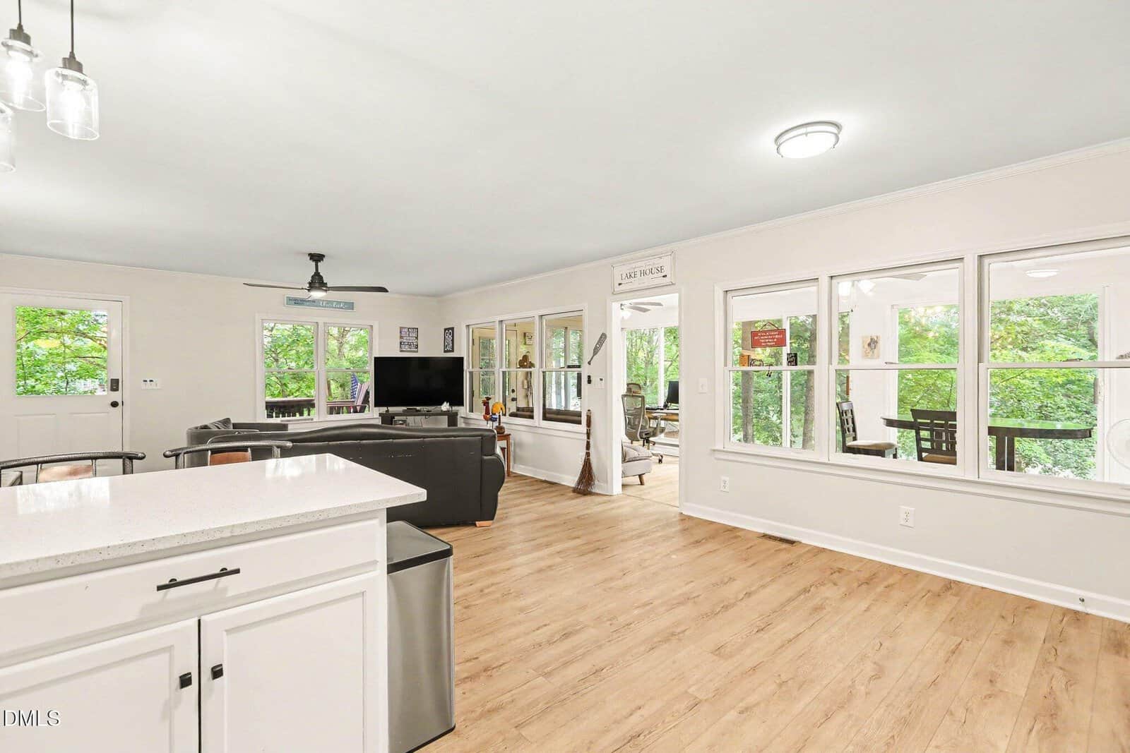 Bright open living area with wood flooring, white walls, and large windows. Modern kitchen with white cabinets, living room with black couch and TV, and a sunroom visible through French doors. Green trees outside.