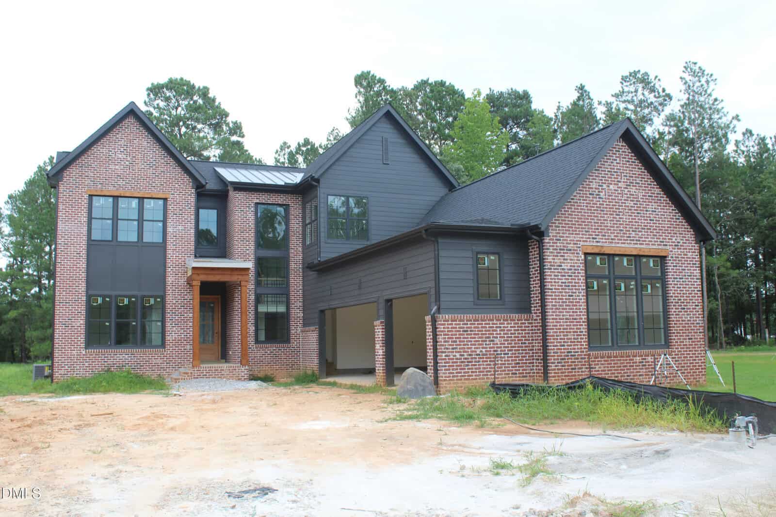 Two-story modern house at 10 Geranium Drive, Youngsville, featuring a mix of red brick and dark siding exterior, large windows, and a sloped roof. The yard is unlandscaped with patches of dirt and grass. Pine trees are visible in the background.