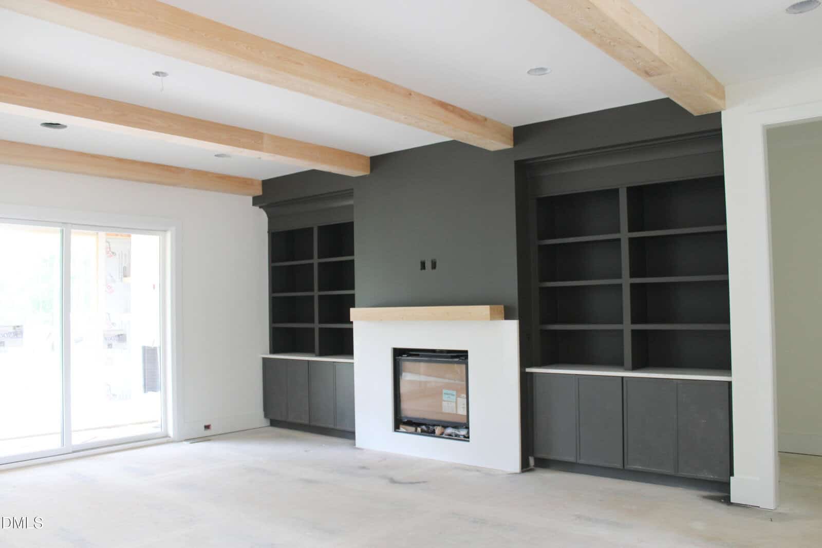 Modern living room with exposed wood ceiling beams, dark built-in shelves on both sides of a fireplace with a light wood mantel, and large sliding glass doors letting in natural light.