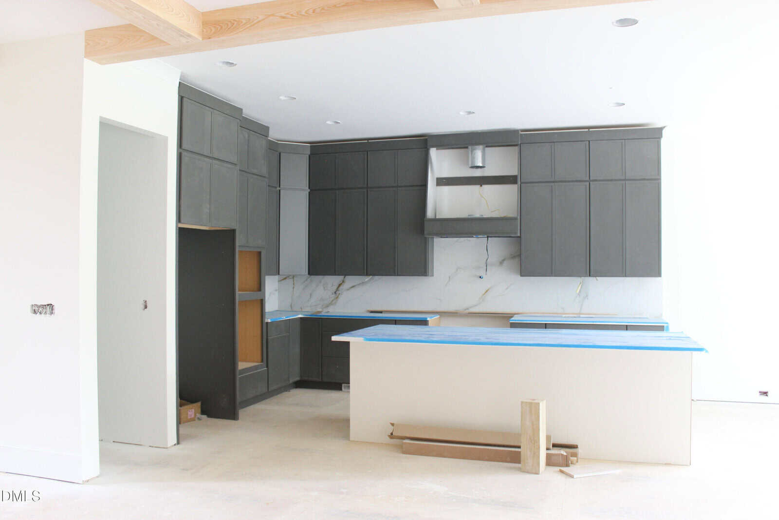 A modern kitchen under construction with dark gray cabinets, white marble backsplash, blue protective wrap on countertops, an island in the center, and unfinished floors.