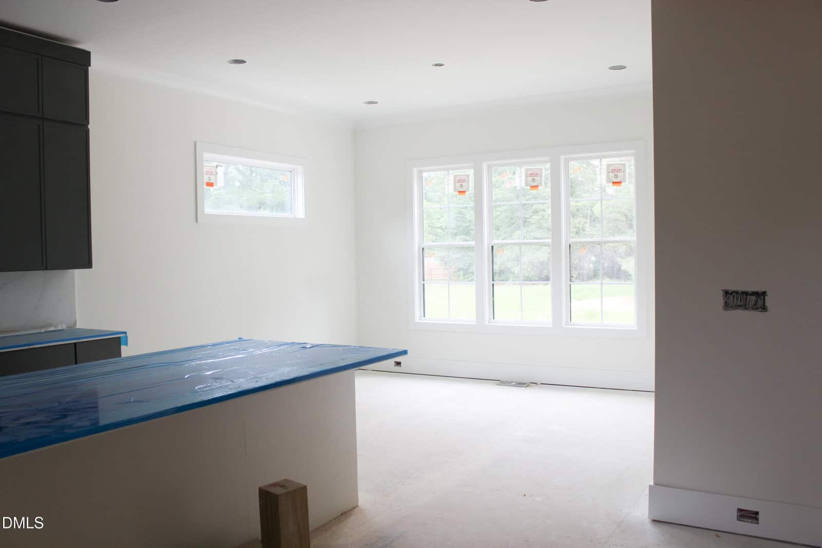 An unfinished kitchen with white walls, a covered island countertop, dark cabinets, and large windows allowing natural light into the empty space. The floor appears to be under construction.
