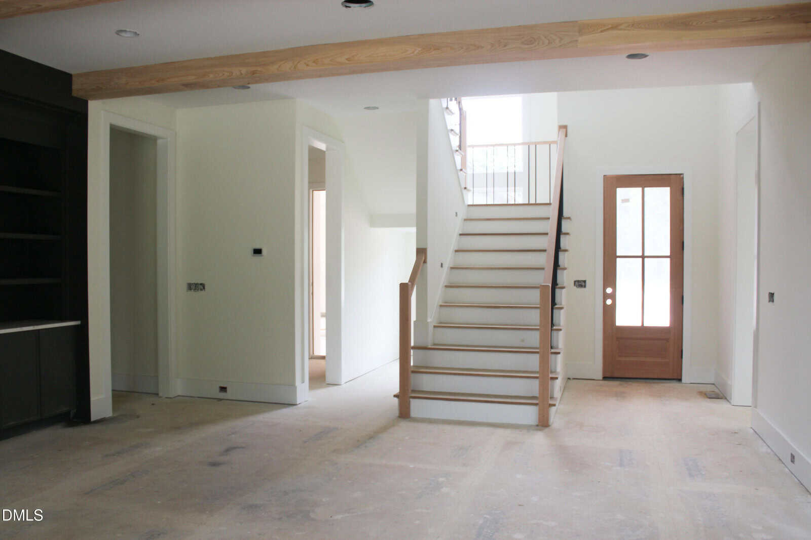 Unfinished interior of a modern home with a wooden staircase, white walls, exposed ceiling beams, an empty floor, a wooden door with glass panels, and natural light coming from a window above the stairs.