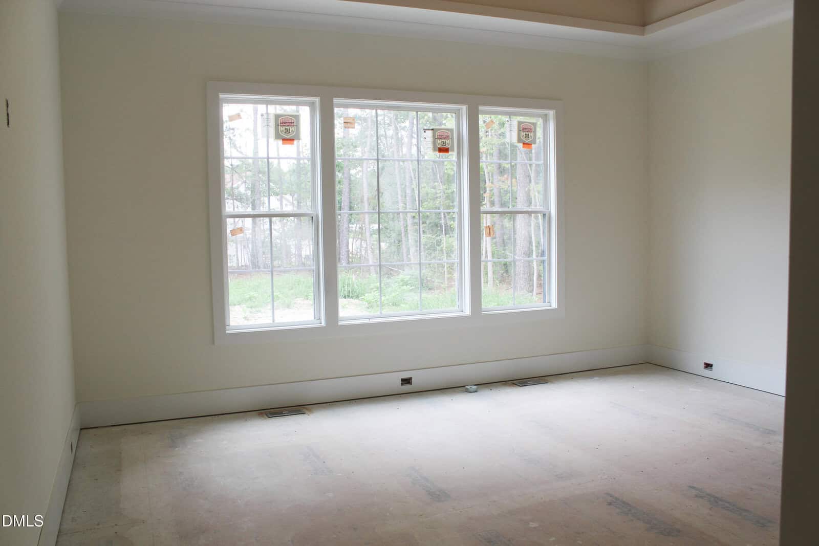 An empty, unfurnished room with off-white walls, three large windows letting in natural light, and an unfinished floor. Trees and greenery are visible outside the windows.