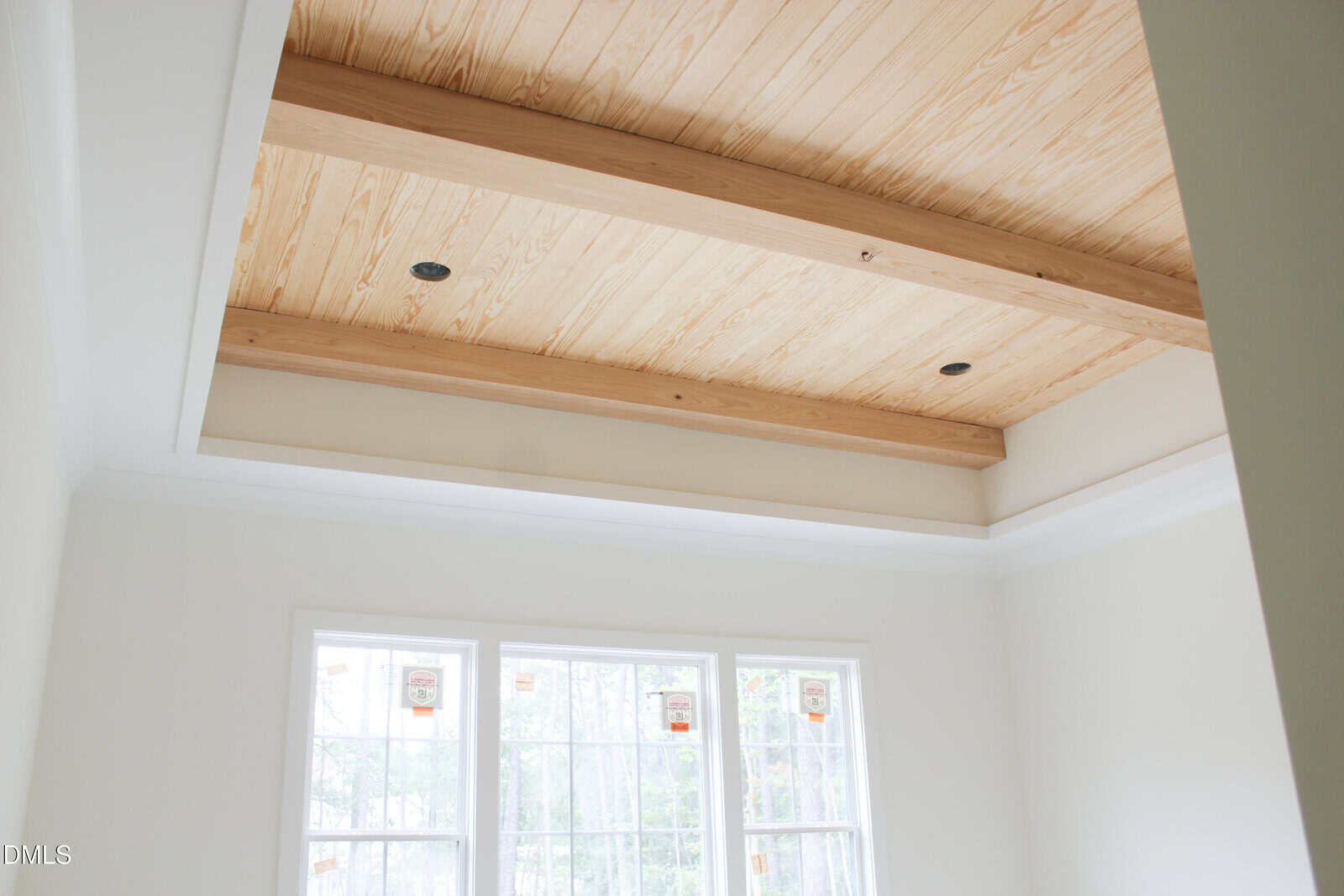 A room with large windows and a vaulted ceiling featuring exposed wooden beams and light wood paneling. The walls are painted white, and natural light fills the space.