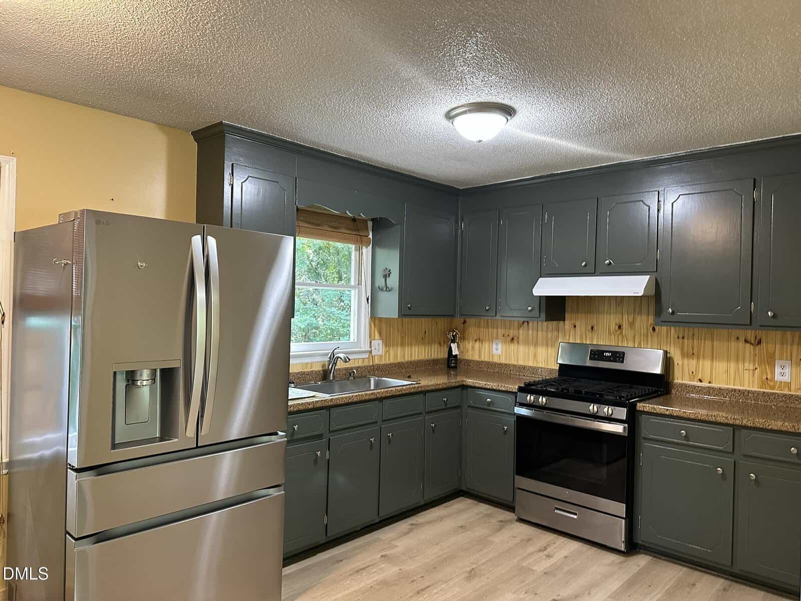 Modern kitchen with gray cabinets, stainless steel refrigerator, gas stove, and vent hood. There is a window above the sink, light wood floors, tan backsplash, and overhead lighting.