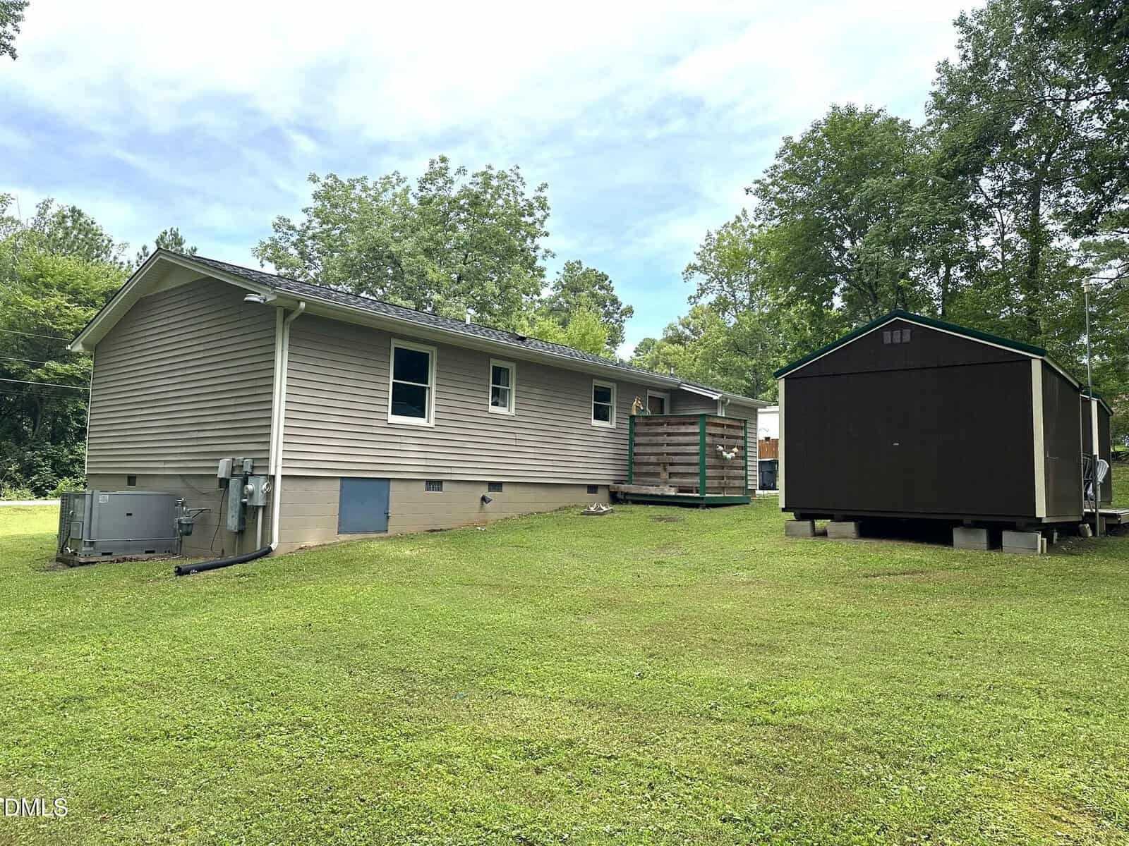 A beige single-story house with a small deck sits on a grassy lawn. A dark brown shed is nearby. Lush green trees surround the backyard, and the sky is lightly cloudy.