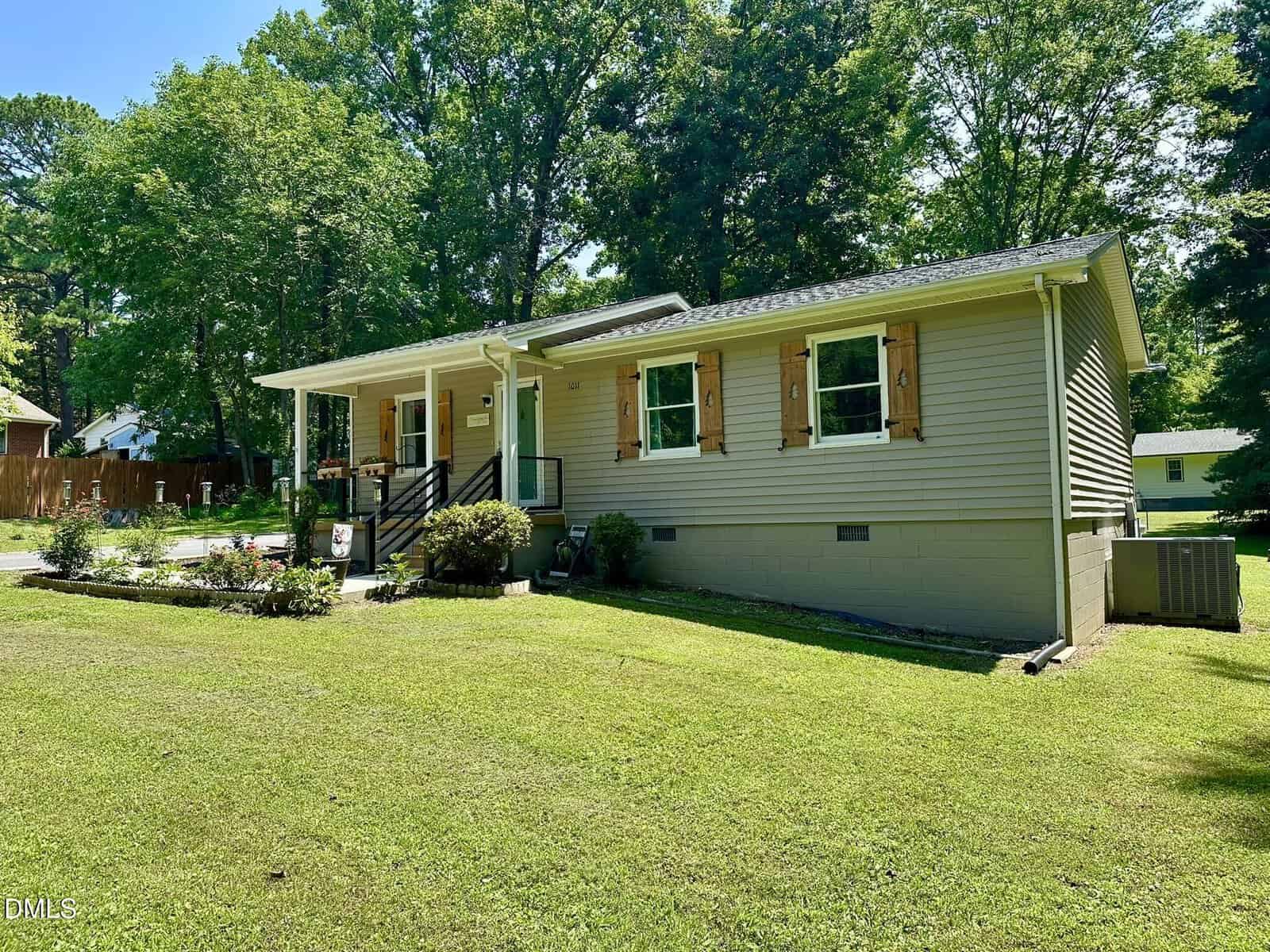 Single-story beige house with a front porch, wooden shutters, and a well-kept lawn. There are shrubs along the foundation, tall trees in the background, and a driveway to the left. The scene is sunny with clear skies.