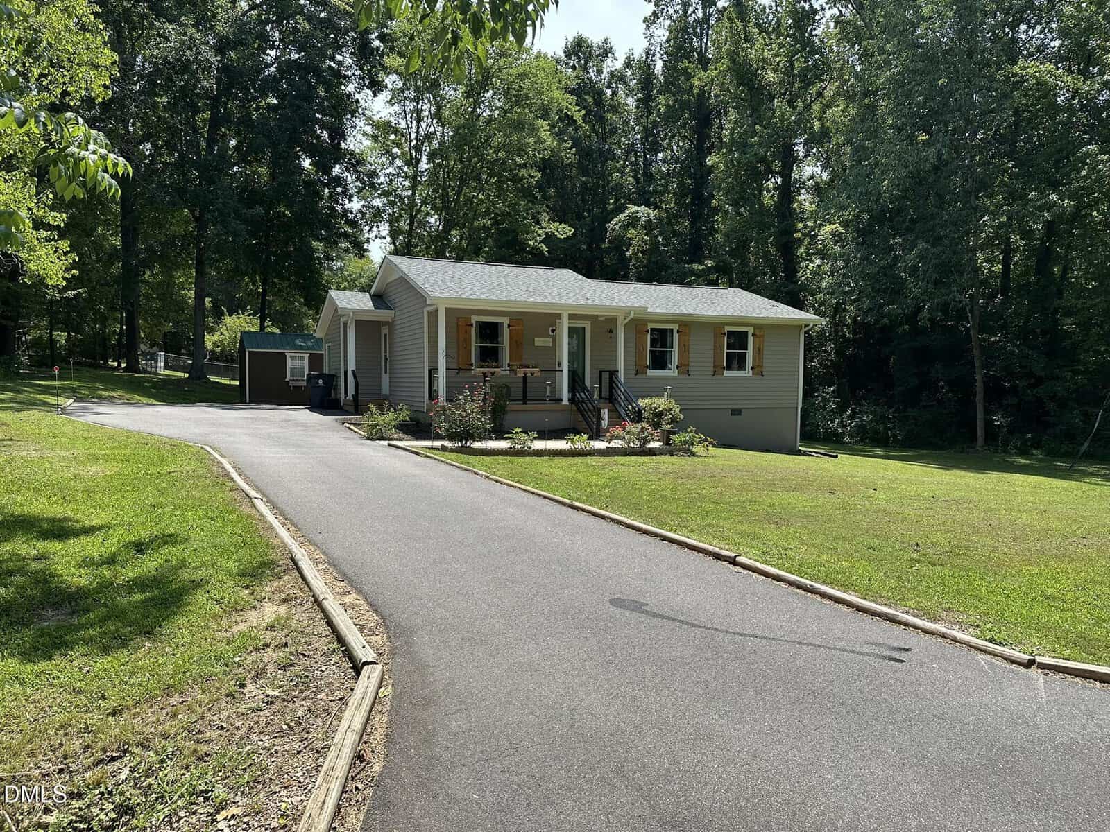 A single-story beige house with a front porch and shutters sits at the end of a paved driveway, surrounded by a grassy lawn and dense trees in the background. A small shed is visible to the left of the house.