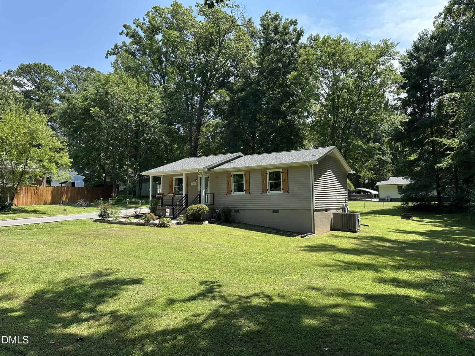 A single-story house with beige siding and white trim sits on a large, well-kept lawn. Mature trees surround the property, and a wooden fence is visible on the left side. The sky is clear and sunny.