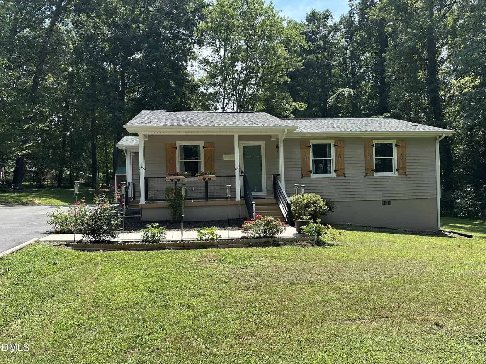 Single-story house with gray siding, wooden shutters, a front porch with a ramp, and flower beds, surrounded by green grass and trees on a sunny day.