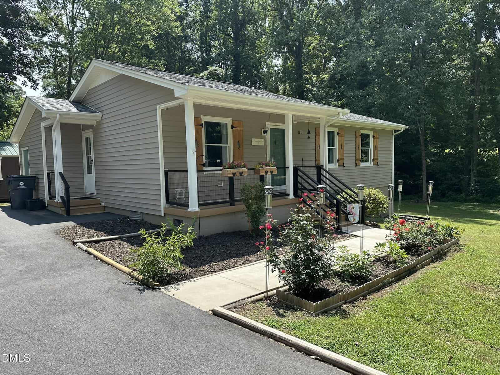 Single-story beige house with a small front porch, black railings, and flower boxes. A landscaped garden with blooming flowers lines the walkway. Surrounded by trees and green grass, the paved driveway leads to the entrance.