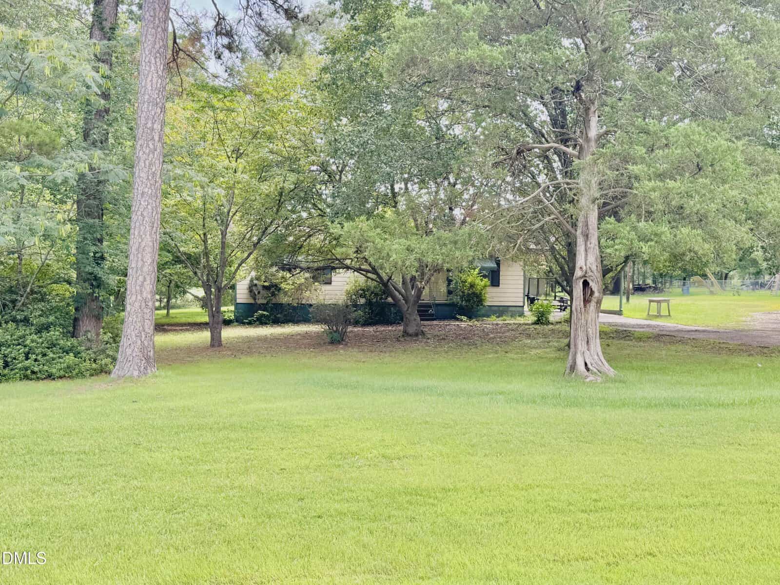 A small light-colored house is partially visible behind trees, surrounded by a large green lawn and shaded by tall trees on a bright day.