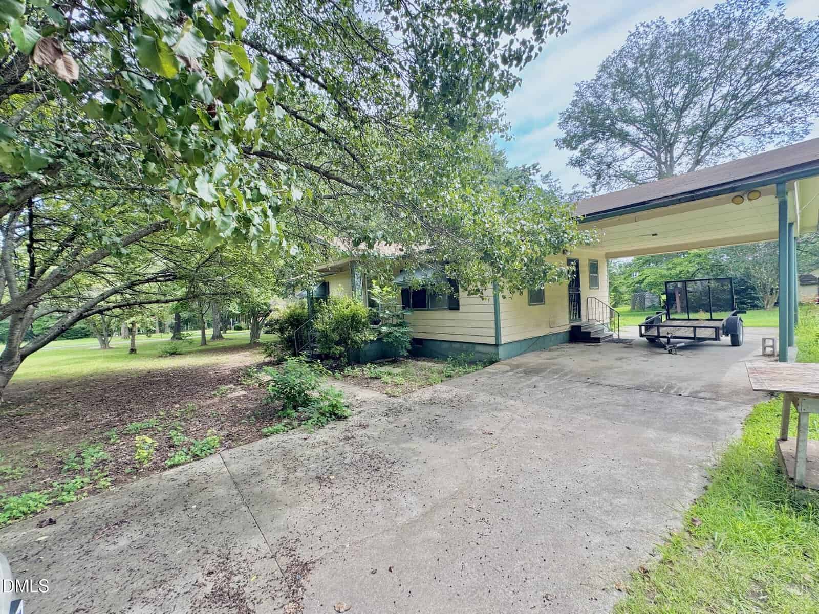 Single-story yellow house with a covered carport, concrete driveway, and large trees providing shade around the property. A utility trailer is parked under the carport. Grass and greenery are visible throughout the yard.