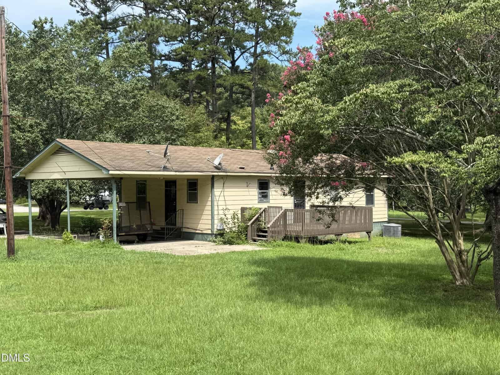 Single-story yellow house with tan roof, front porch, ramp, and surrounded by green grass, trees, and shrubs on a sunny day.