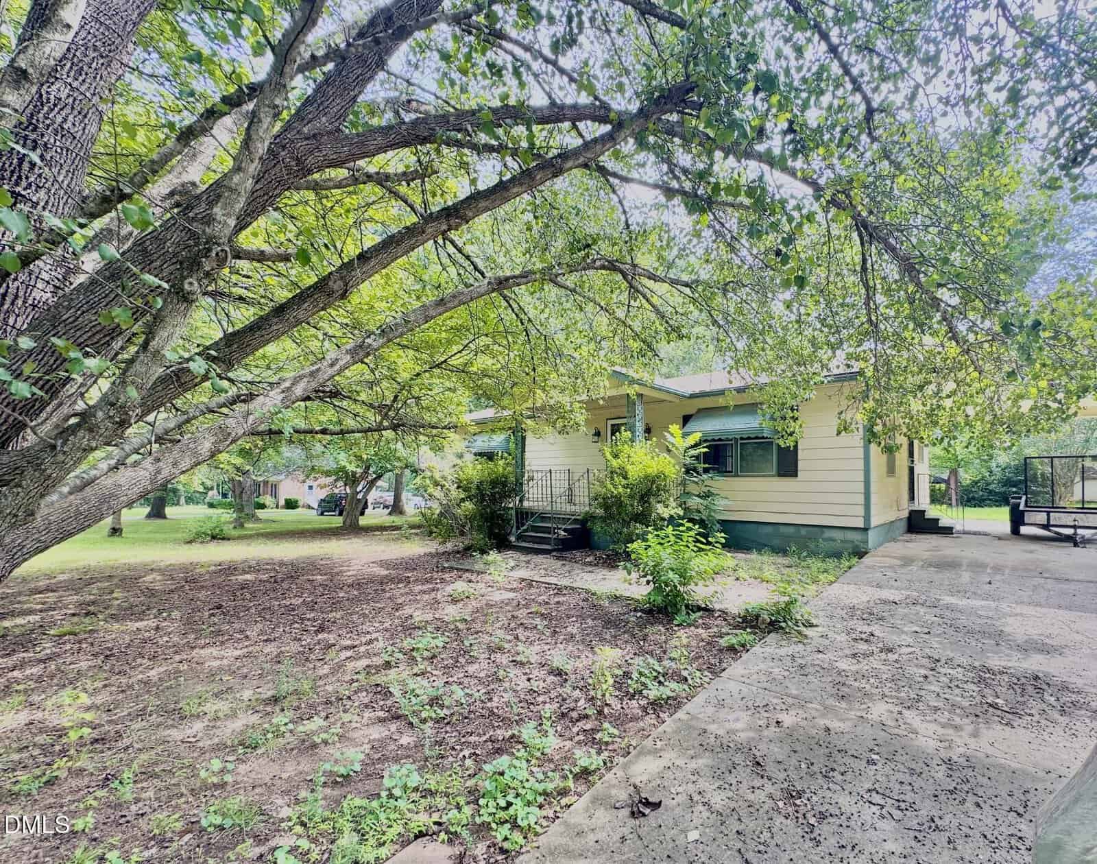 Single-story yellow house with green shutters, surrounded by large trees and greenery. A concrete driveway leads to the front steps and porch area. The yard is mostly shaded with patches of sunlight on the ground.