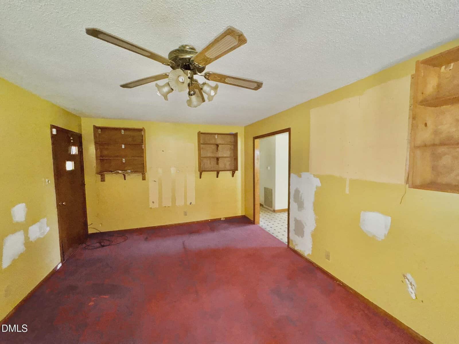 Empty room with yellow walls, red carpet, wooden shelves, missing wall fixtures, a ceiling fan with lights, and visible patches on the walls. An open doorway leads to a room with a white tiled floor.
